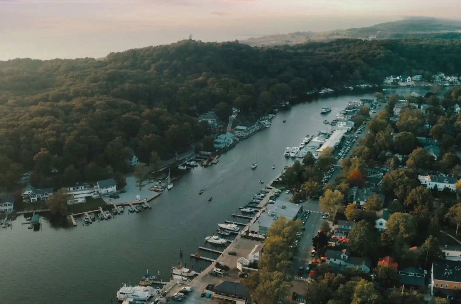 Aerial view of a marina with boats docked along a river flanked by dense forest and residential houses.