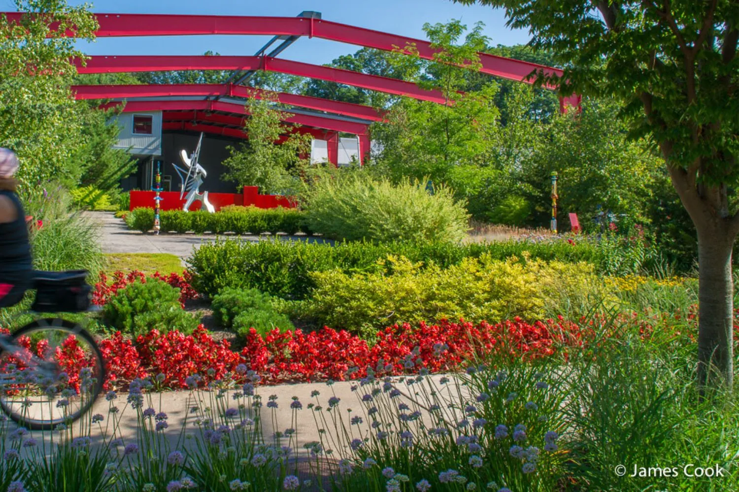Colorful garden with red flowers, green bushes, and a cyclist passing on a path near a structure with red beams and a metal sculpture.