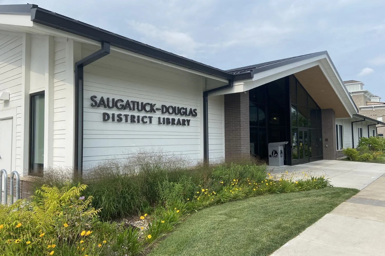 Exterior view of Saugatuck-Douglas District Library with white siding, dark trim, large windows, and landscaping with yellow flowers.