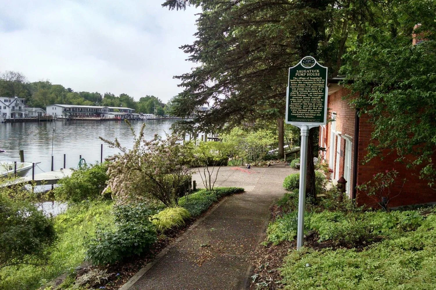 Pathway beside a brick building with greenery leading to a water body with docks and buildings across, featuring a historical plaque about the Saugatuck Pump House.