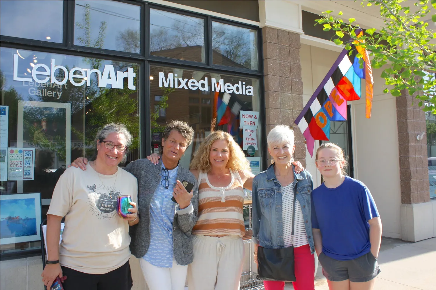 Five smiling people standing arm in arm outside LebenArt Mixed Media gallery with an open flag visible.