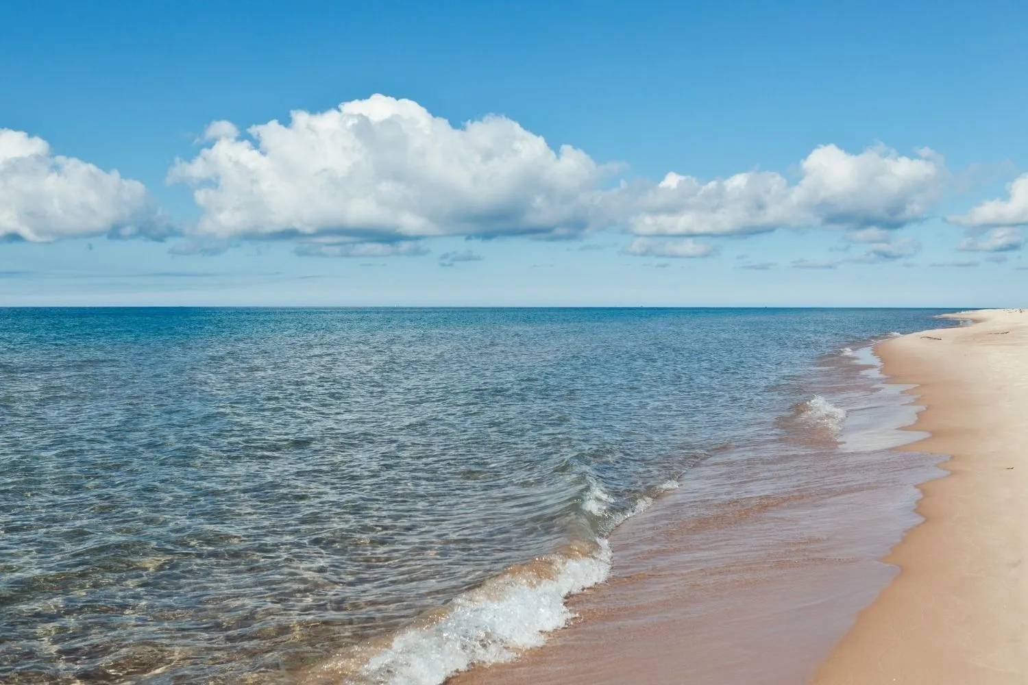 Clear shallow waves gently lapping onto a sandy beach under a blue sky with scattered white clouds.