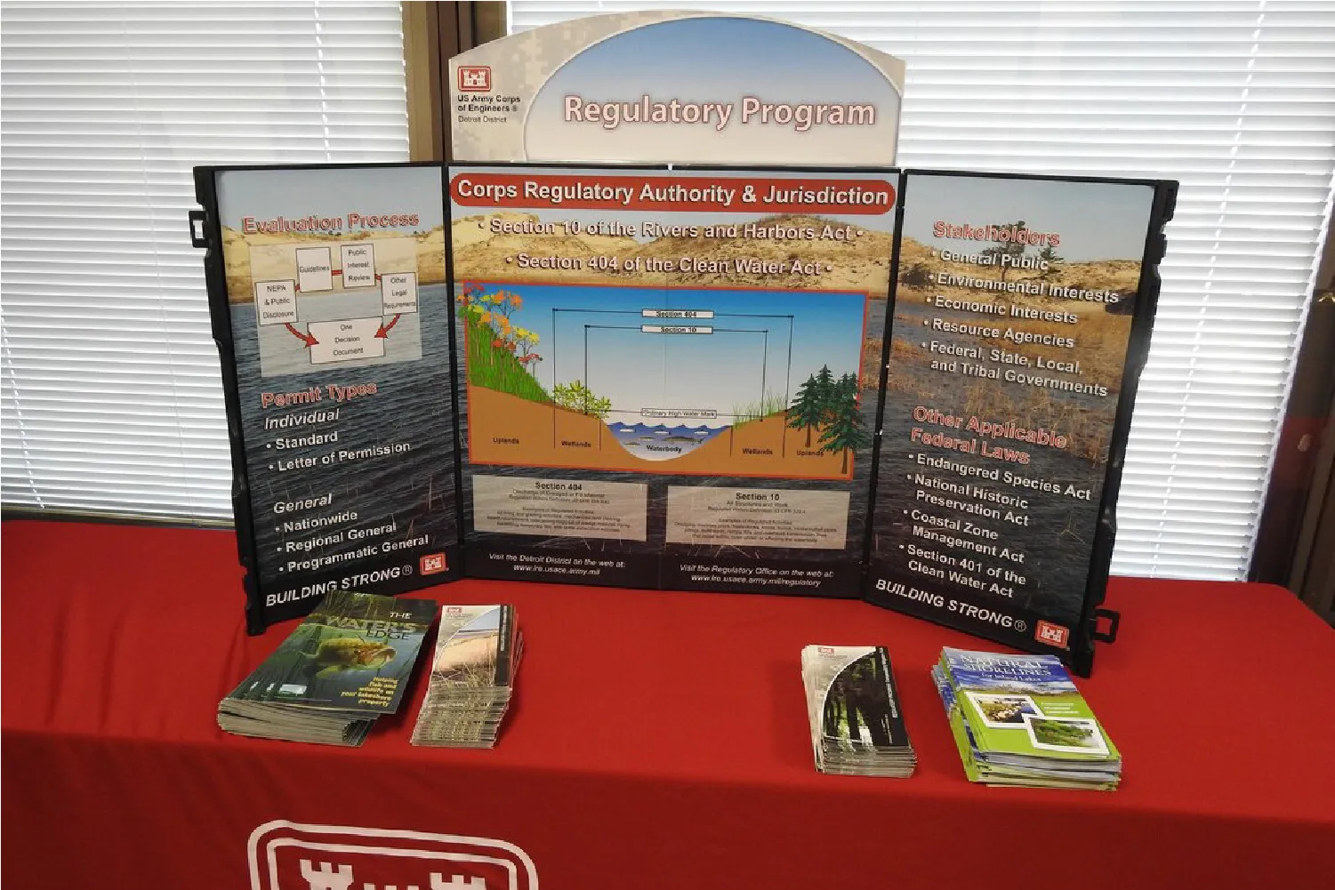 Three-panel informational display about US Army Corps of Engineers Regulatory Program on a red table with brochures.