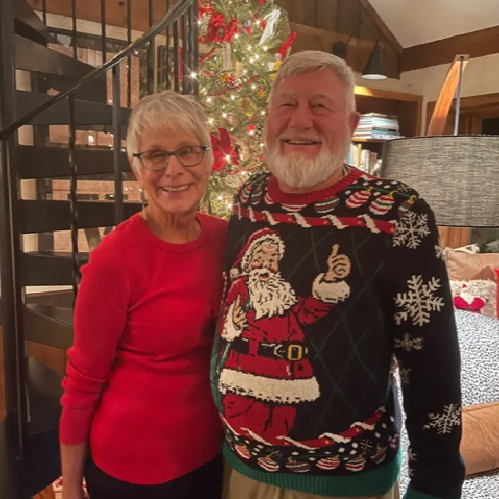 Smiling elderly couple standing indoors in front of a decorated Christmas tree, the man wearing a Santa-themed holiday sweater and the woman in a red sweater.