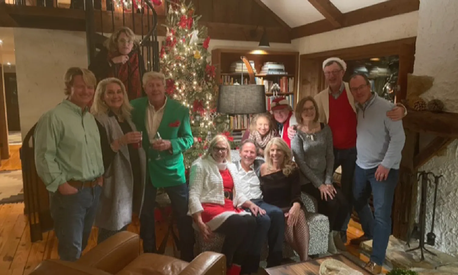 Group of twelve people posing indoors around a decorated Christmas tree in a cozy living room with festive attire and warm lighting.