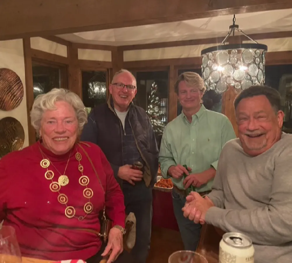 Four adults smiling and posing in a cozy room with wooden beams and a decorative lamp, with a Christmas tree visible through the window behind them.