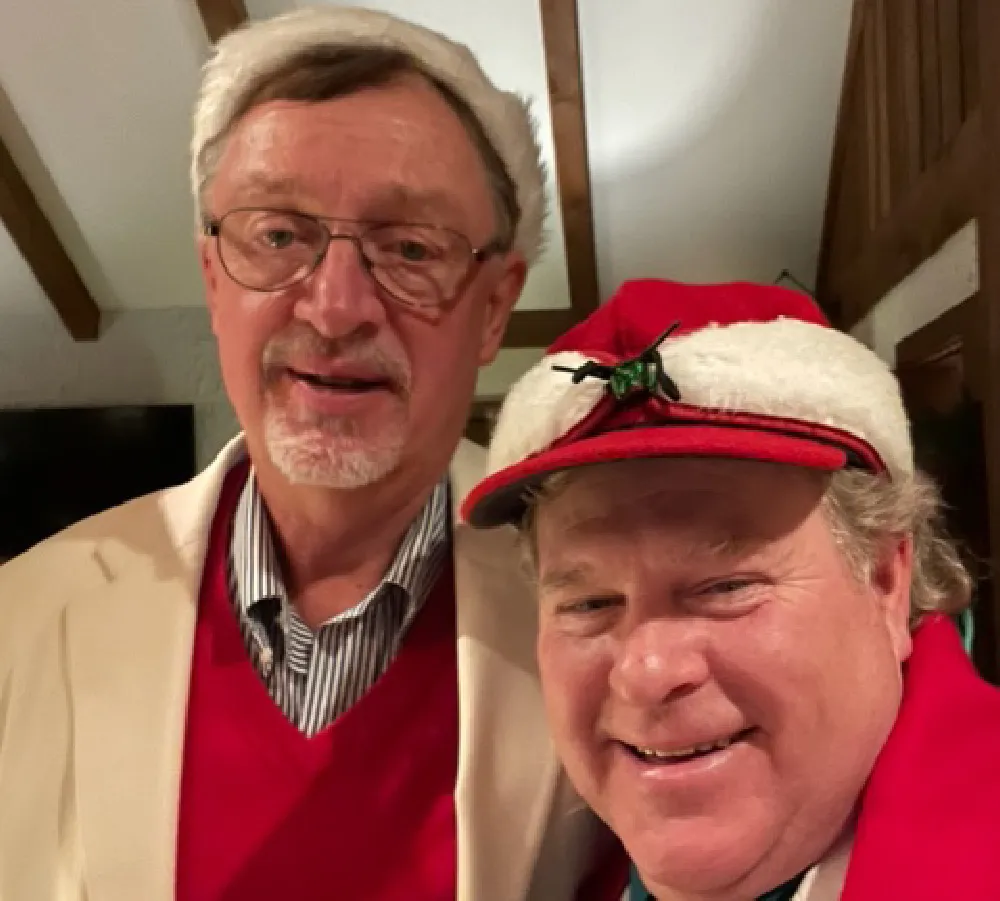 Two men wearing Santa hats and festive attire smiling indoors under wooden ceiling beams.