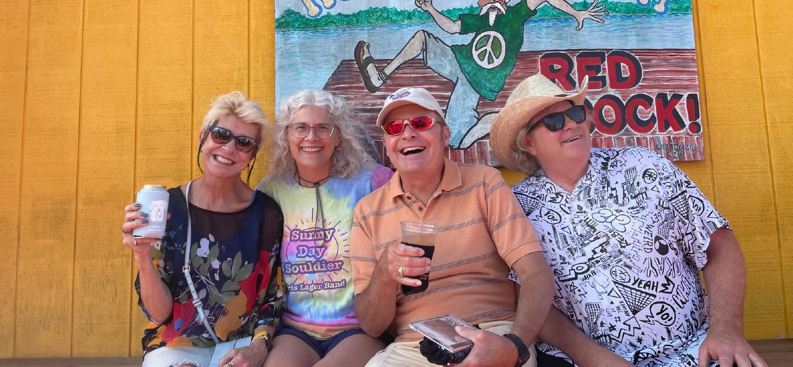 Four smiling adults sitting on a bench with drinks, in front of a yellow wall and a colorful mural.