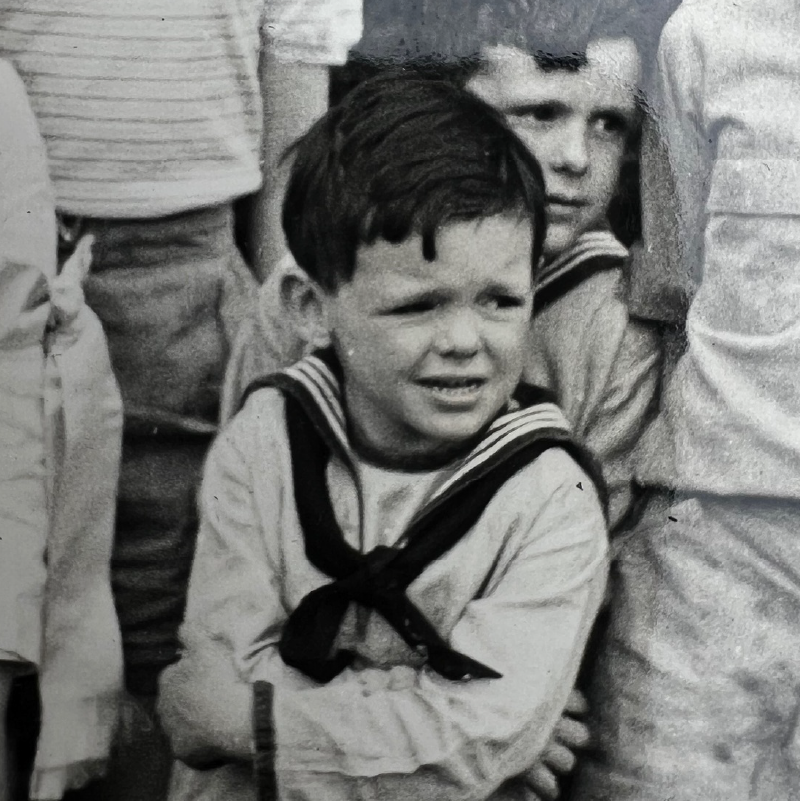 Black and white close-up of a young boy wearing a sailor-style outfit, looking to the side with a concerned expression.