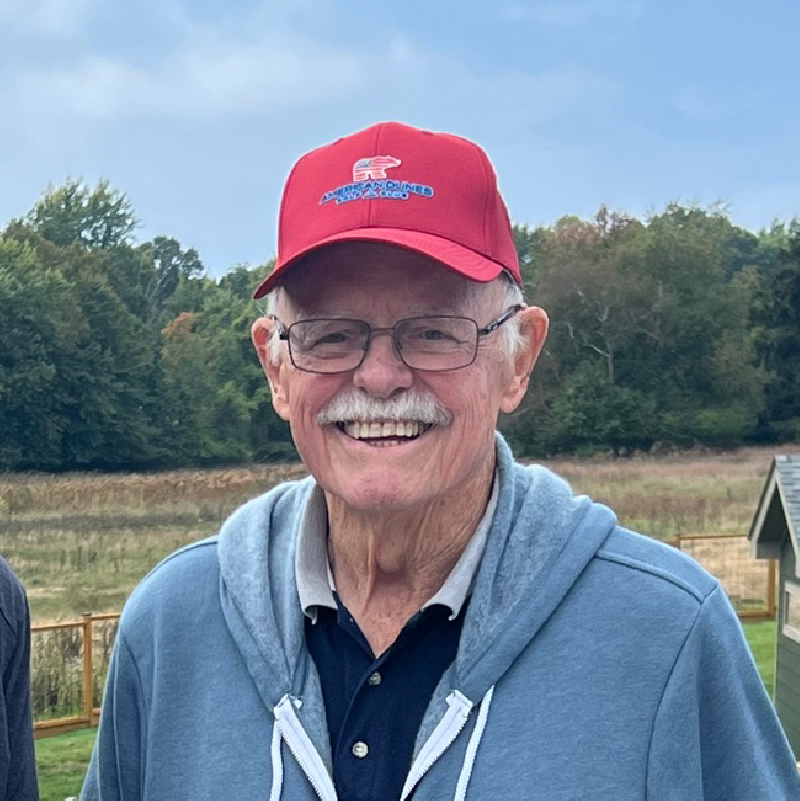 Smiling elderly man wearing glasses, a red cap, and a blue hoodie standing outdoors with a field and trees in the background.