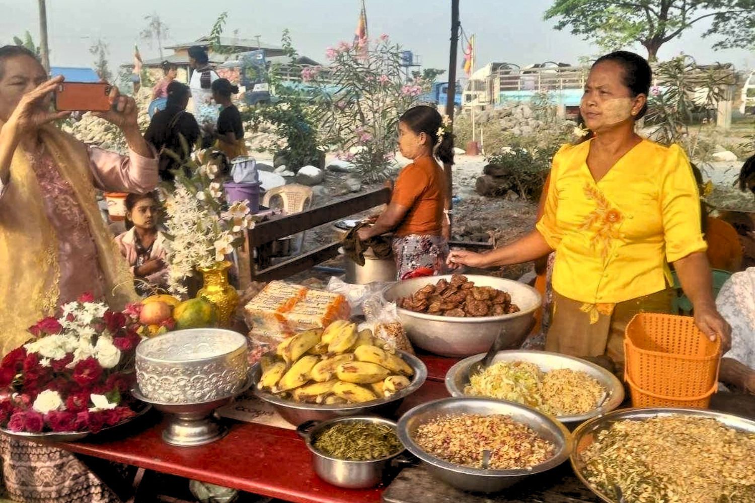 A woman in a bright yellow blouse with thanaka on her cheeks stands beside a table spread with traditional Myanmar dishes, fresh bananas, and floral displays in Kalay Township.