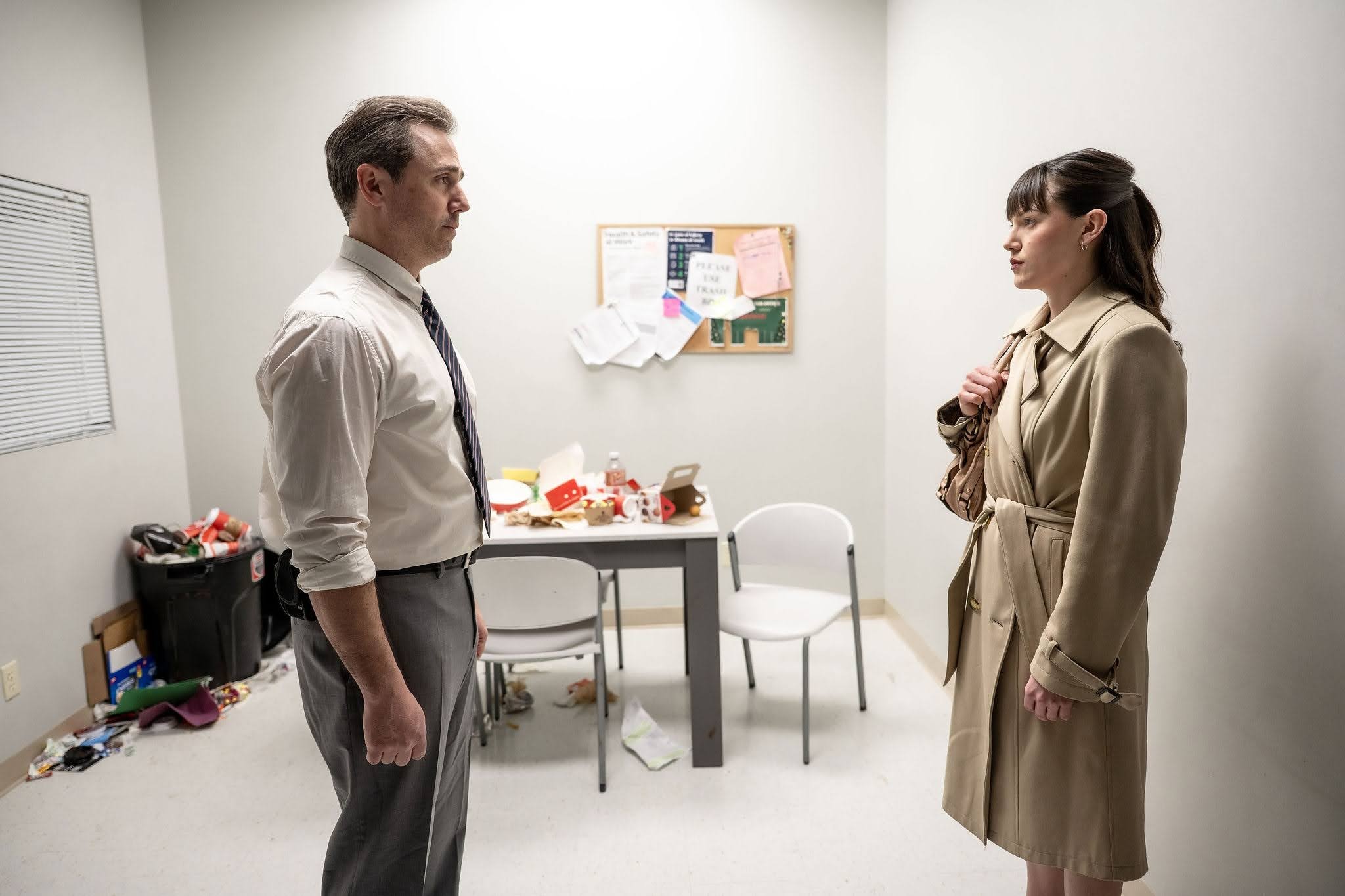 A middle-aged white man with salt and pepper hair wearing a white shirt, dark tie and grey pants stands facing a white woman in her twenties with long brown hair and a trench coat. They are standing in a small office with a table covered in food wrappers and other garbage one might find in an office break room. 