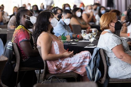 A diverse group of people wearing masks sit at round tables during the NPS 2022 gathering, attentively listening to a speaker.
