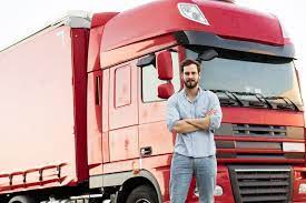 A truck driver wearing light blue long sleeves while arms are crossed in front of a red truck