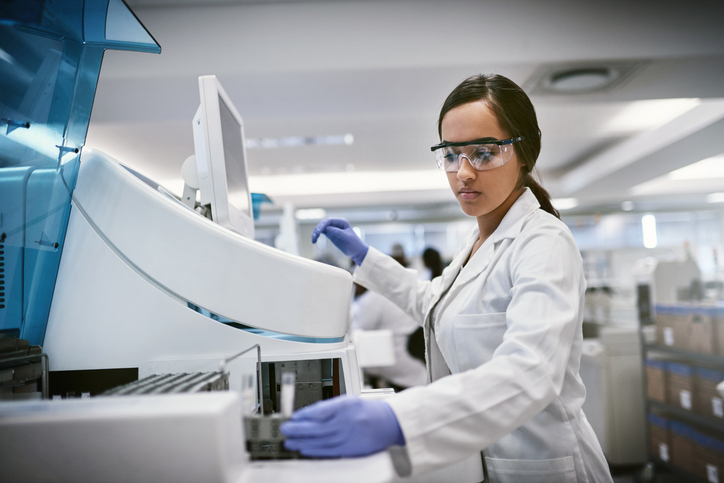 The Medical Technologist is wearing gloves while holding a test tube presumably performing a test and is monitoring the results on a screen