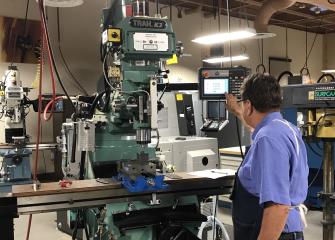 A machinist wearing an apron and glasses is pressing a button on a screen next to a large machine