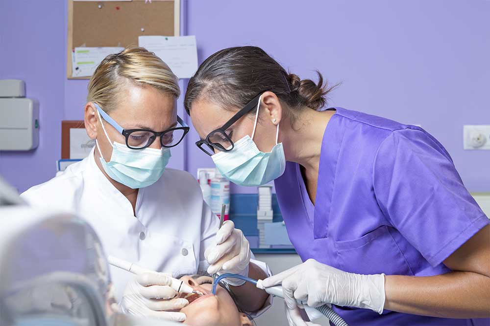 A dental assistant is holding a dental suction device beside a dentist who is cleaning the mouth of a patient