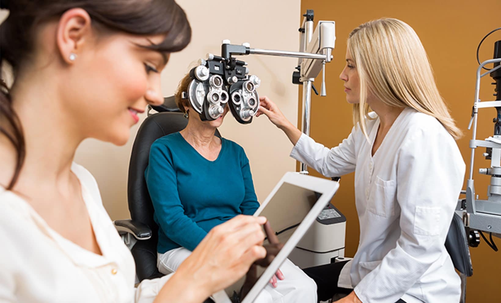 An ophthalmic assistant is reviewing the status of the patient using a tablet while the ophthalmologist performs an eye exam on the patient