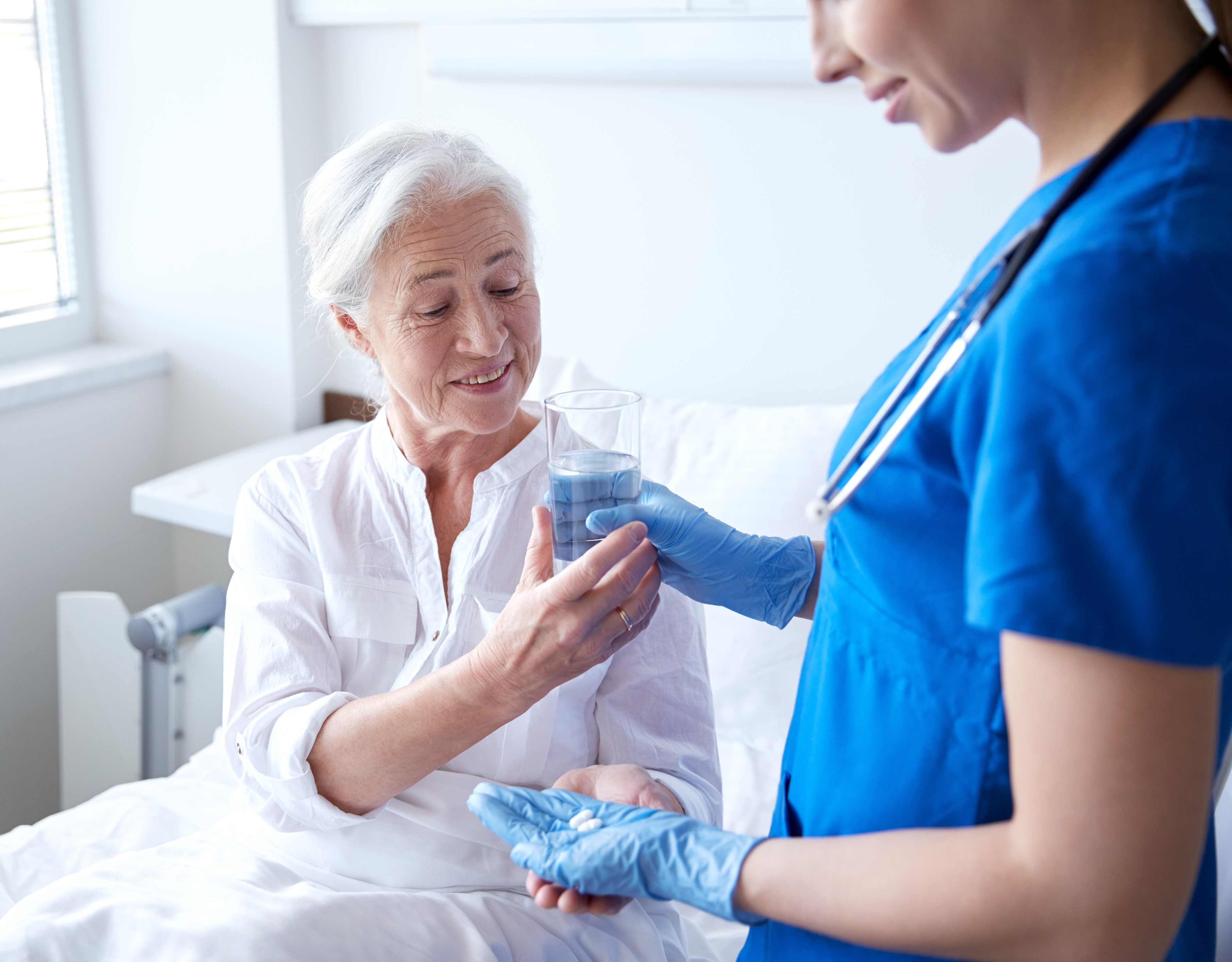 A Medication Aide dressed in scrubs and gloves giving water and medicine to an elderly woman in a hospital bed