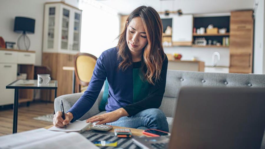 A bookkeeper sitting at her desk with her laptop and calculators, focused on writing in a ledger with a pen
