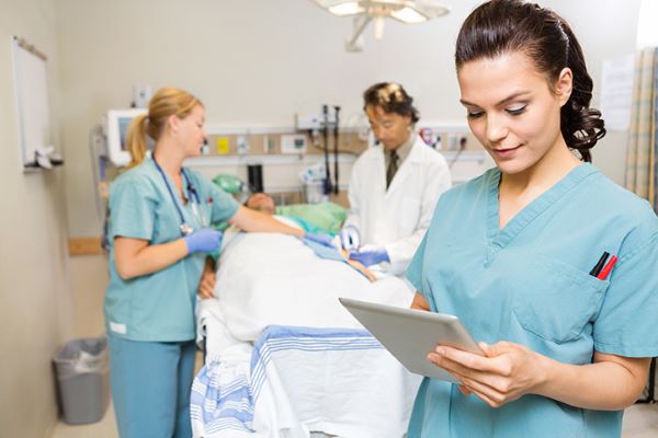 A nurse is holding an iPad while a doctor, another nurse, and a patient are visible in the background