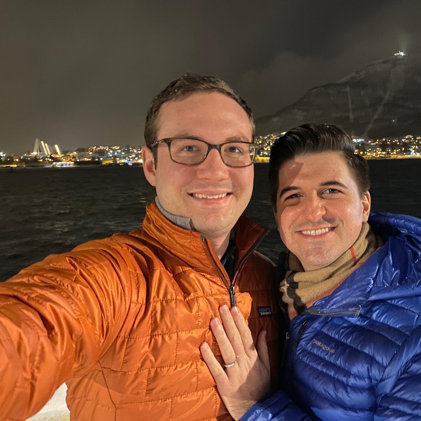 Two men standing in front of the water in Tromsø, Norway. One man has a diamond engagement ring on his finger and holds it to the other man's chest.