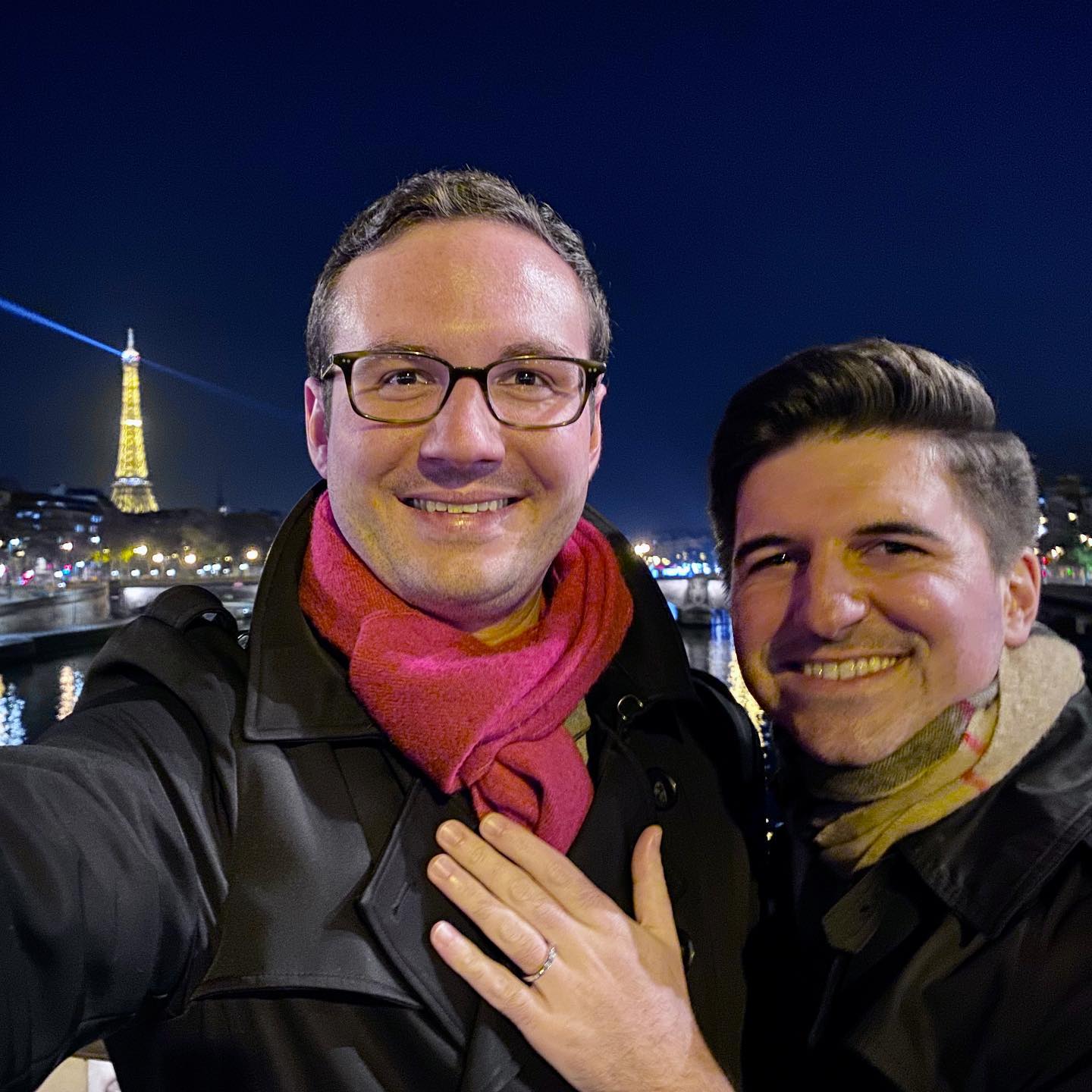 Two men standing in front of the Eiffel Tower in Paris, France. One man has a diamond engagement ring on his finger and holds it to the other man's chest.