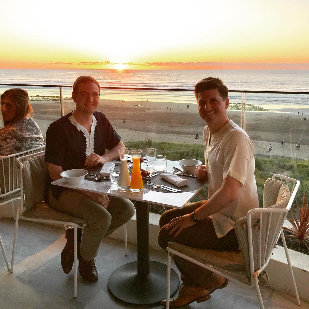 Two men sitting at a table on a balcony overlooking the Atlantic Ocean at sunset.