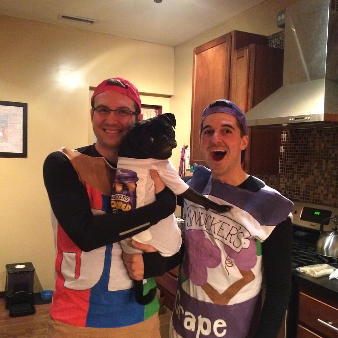 Two men dressed in Halloween costumes -- one of Jif peanut butter, one of Smucker's Grape Jelly -- holding a black pug with a Goober T-shirt on.