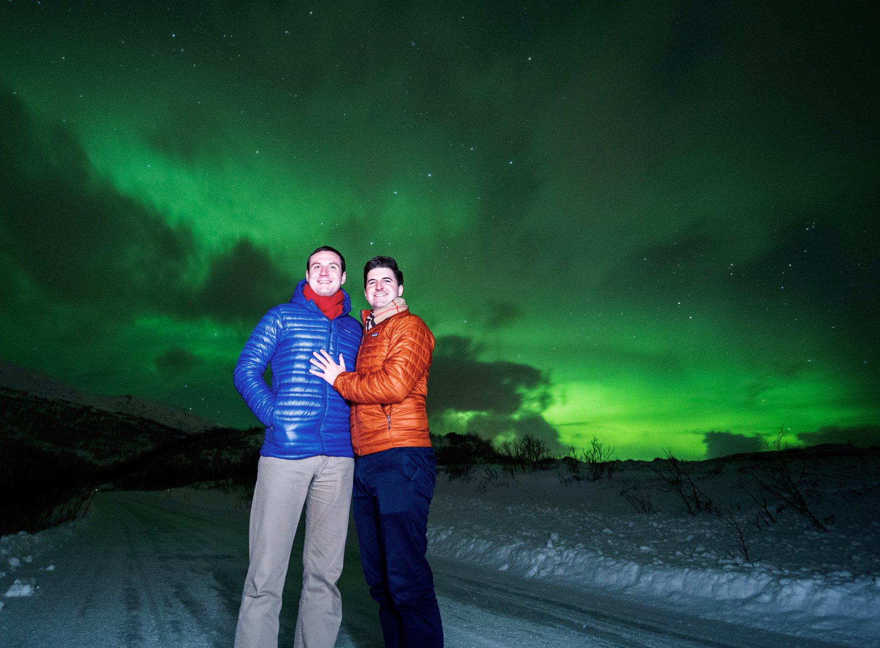 Two men standing in front of the Northern Lights in northern Norway. One man has a diamond engagement ring on his finger and holds it to the other man's chest.