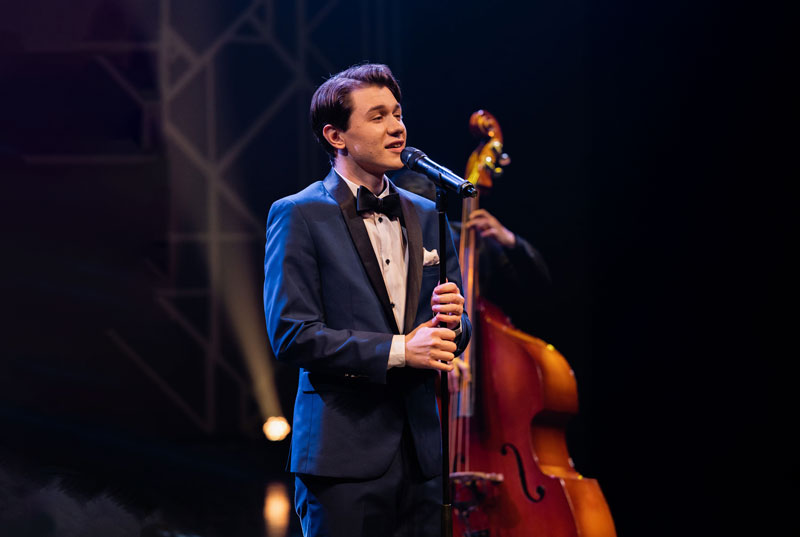 Young male jazz singer in a blue tuxedo performing on stage with a double bass player in the background.