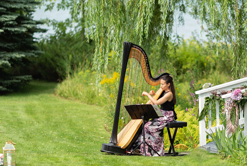 Woman in floral dress playing a harp outdoors under hanging willow branches near decorated white railing.