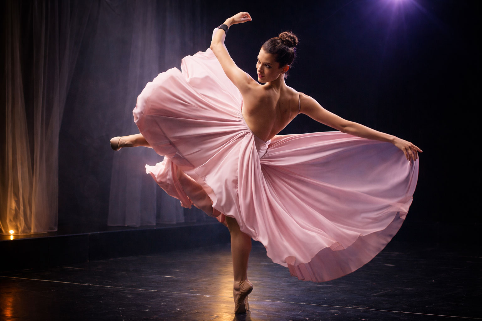 A professional dancer mid-leap in a flowing pink dress performing on a dramatic stage.