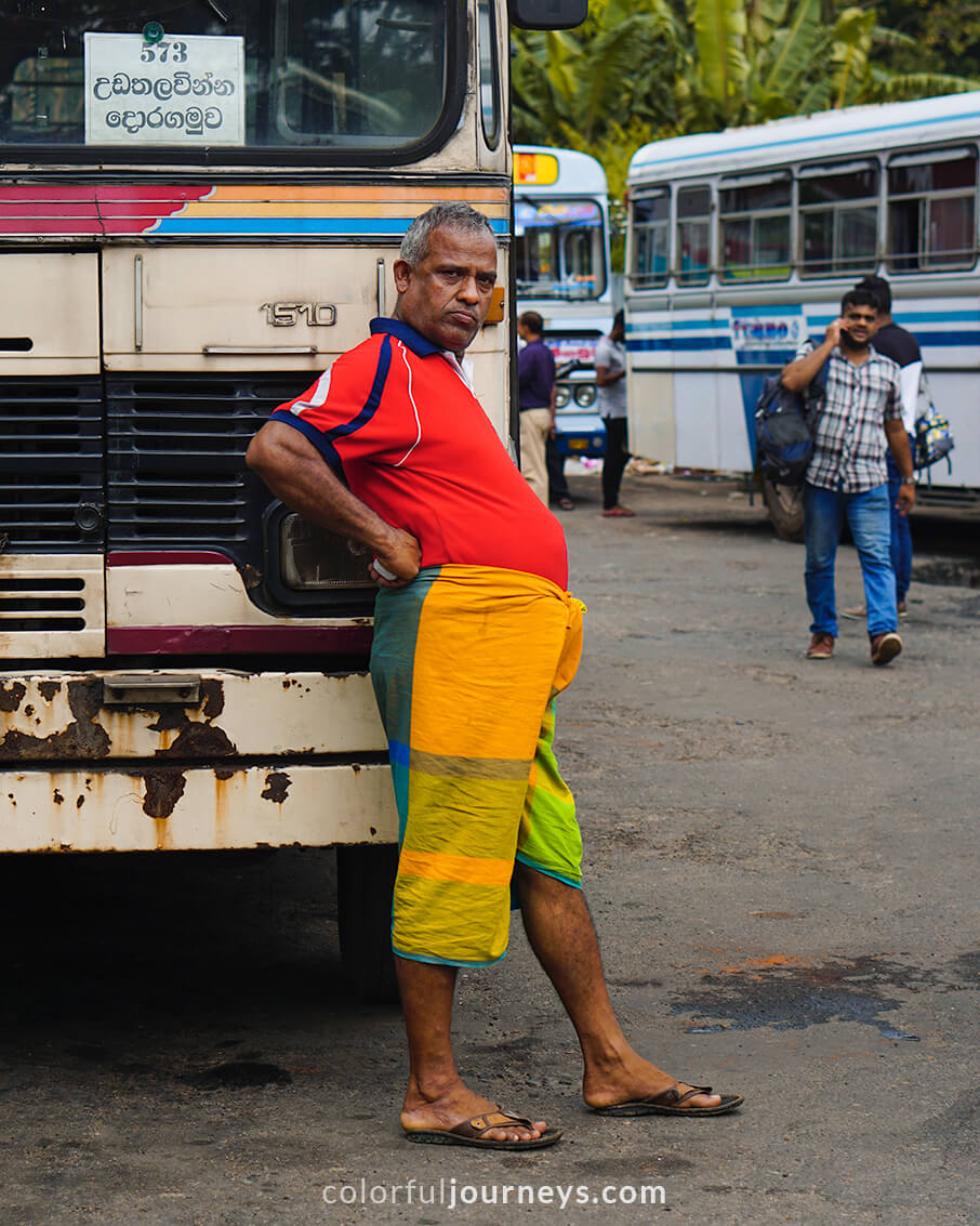 A bus-driver wearing colorful clothes, leans against his bus.