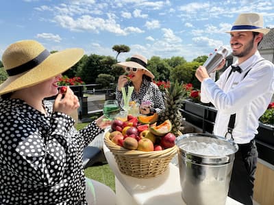 Deux femmes en chapeaux dégustent des fruits, barman prépare un cocktail, ambiance estivale.