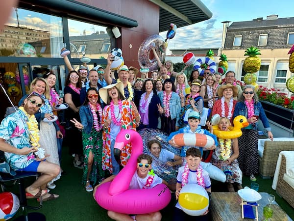 Fête estivale sur rooftop : adultes et enfants souriants, tenues tropicales, bouées colorées.