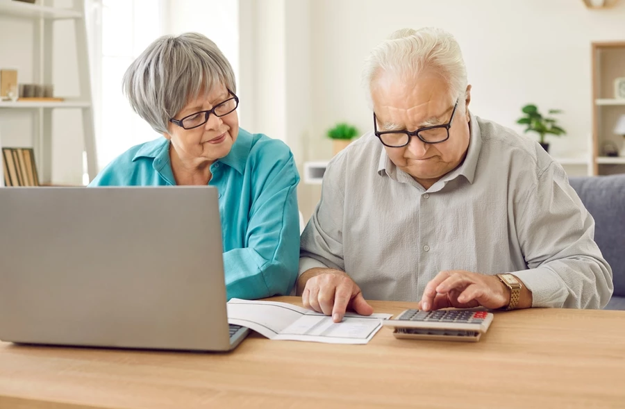 Portrait of elderly couple sitting at home at the desk with laptop and bills calculating finances or taxes.