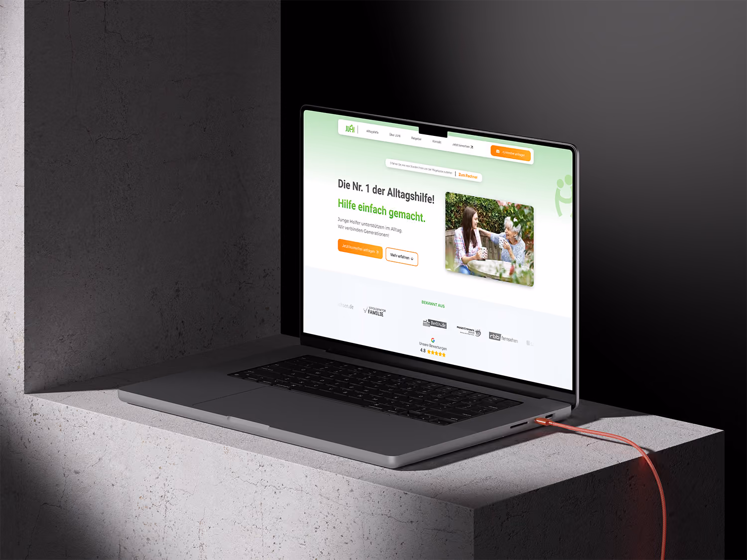 Laptop zeigt Webseite für Alltagshelfer mit Text und Bild von junger Frau mit älterer Frau.