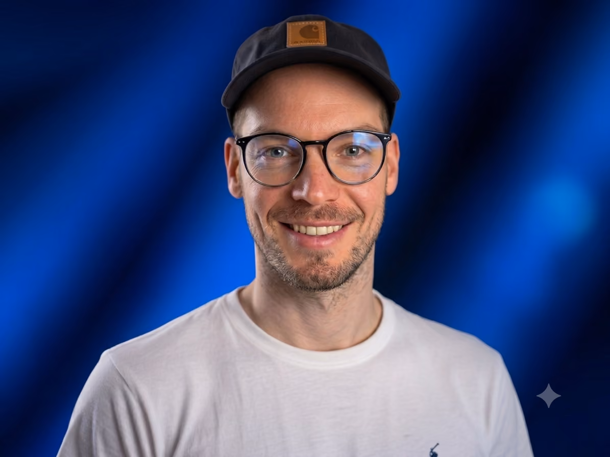 Lächelnder Mann mit Brille, schwarzer Cap und weißem T-Shirt vor blauem Hintergrund.