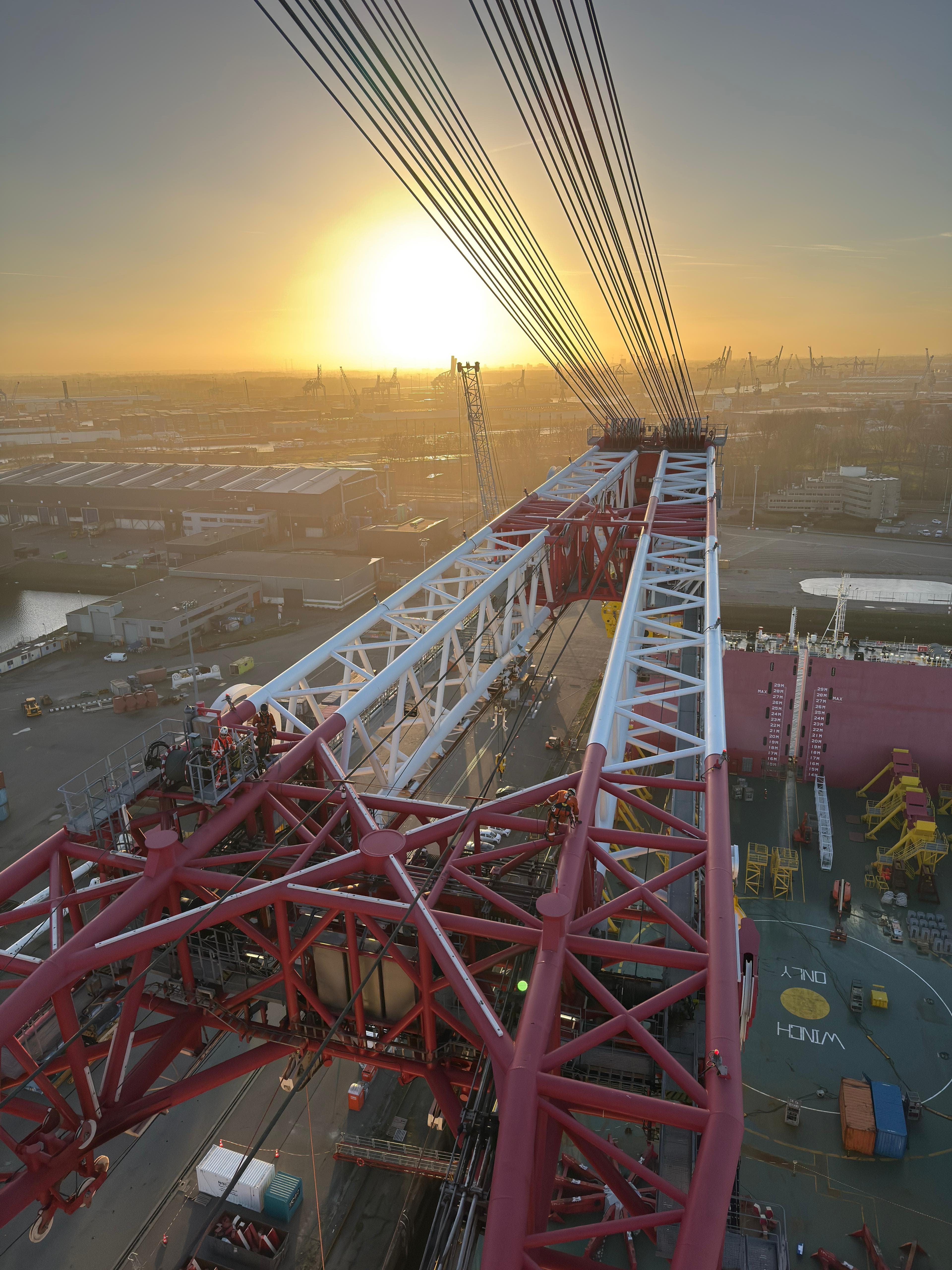 View from top of a large red and white industrial crane at sunset overlooking a port with containers, cranes, and equipment.
