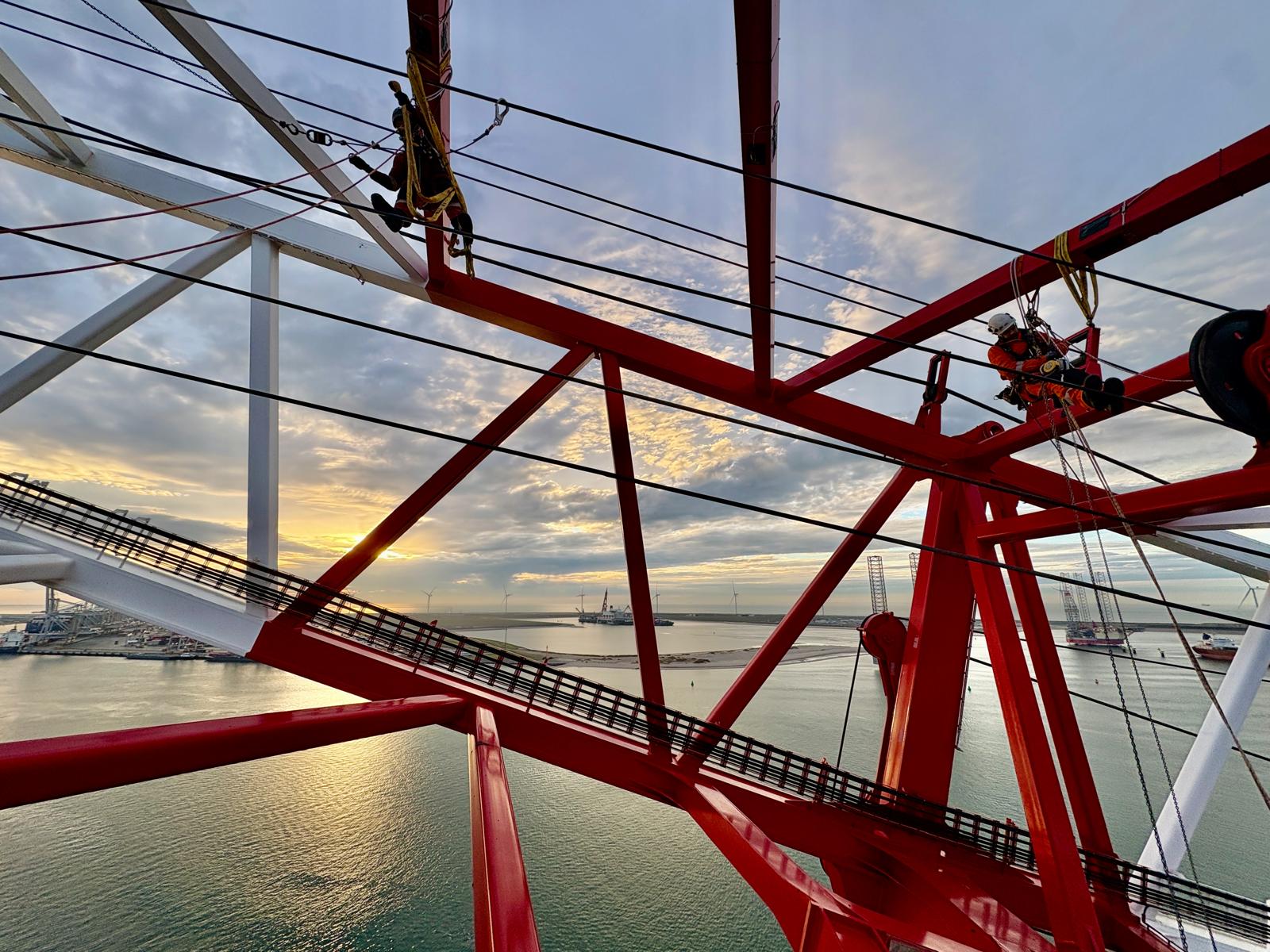 Two industrial workers in safety gear performing rope access maintenance on a large red metal structure above water at sunset.