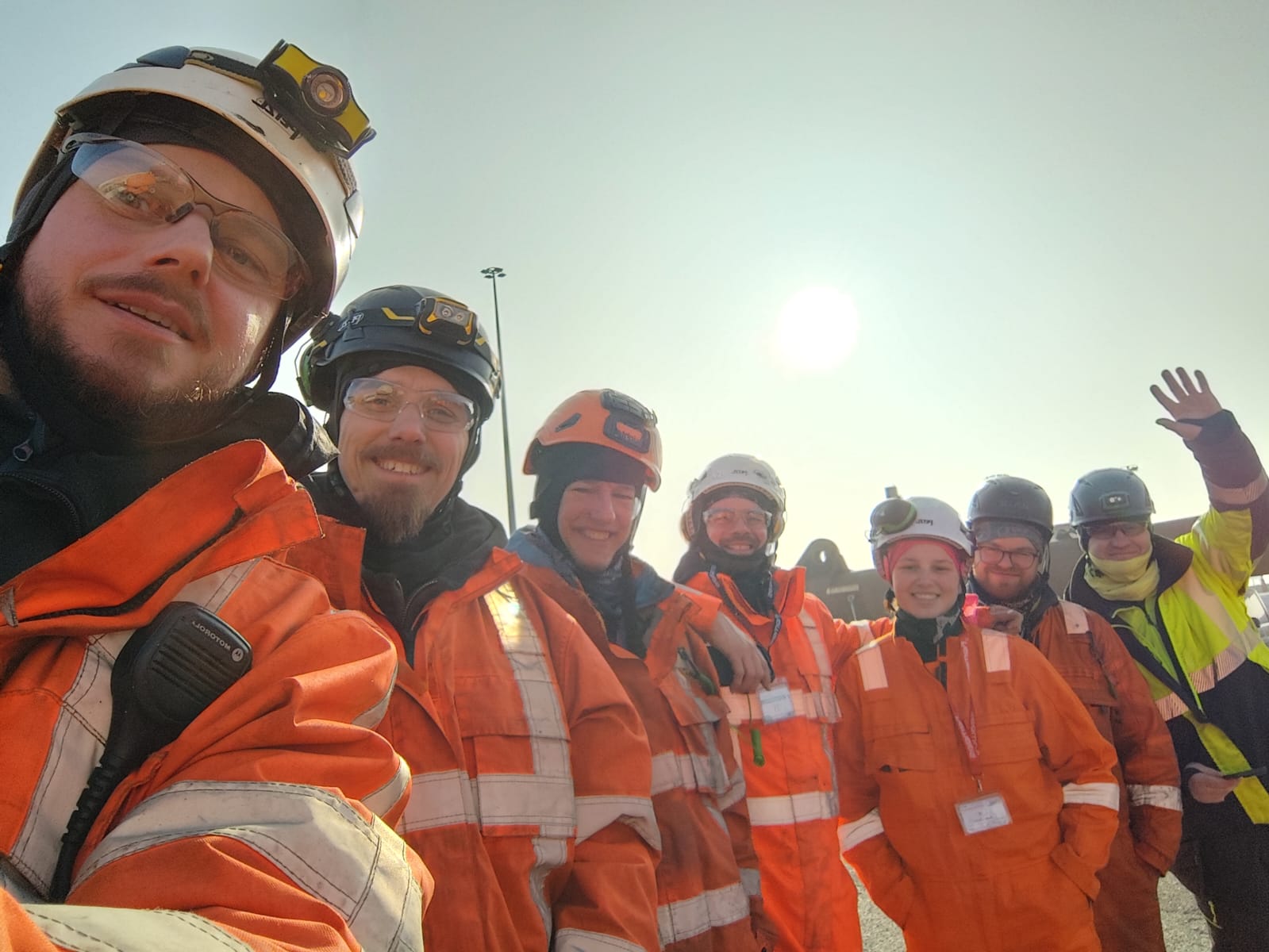 Group of seven workers wearing orange and yellow safety jackets and helmets, smiling at the camera with one waving.
