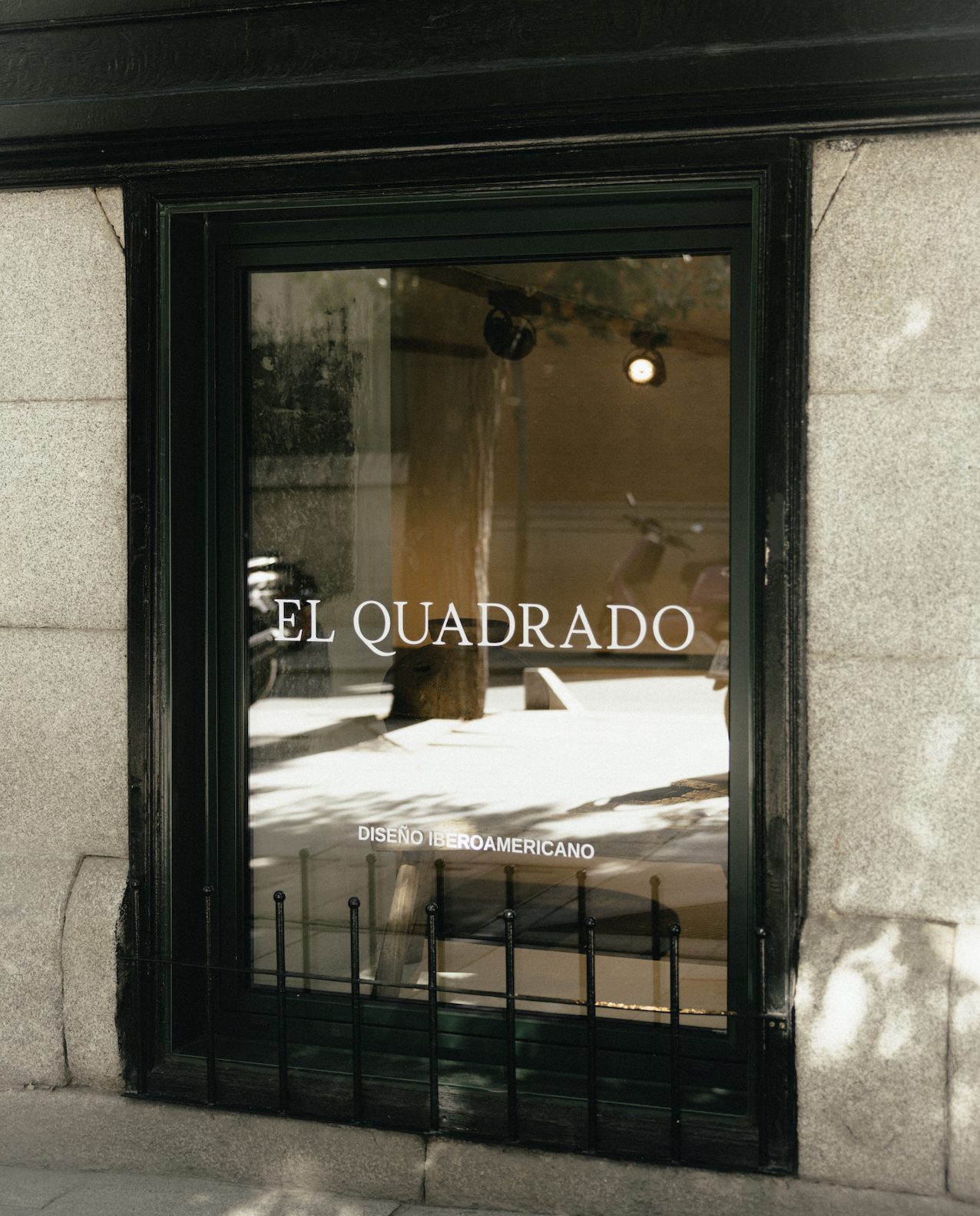Shop window with black frame displaying the text 'EL CUADRADO' and 'DISEÑO IBEROAMERICANO' with street reflections visible.