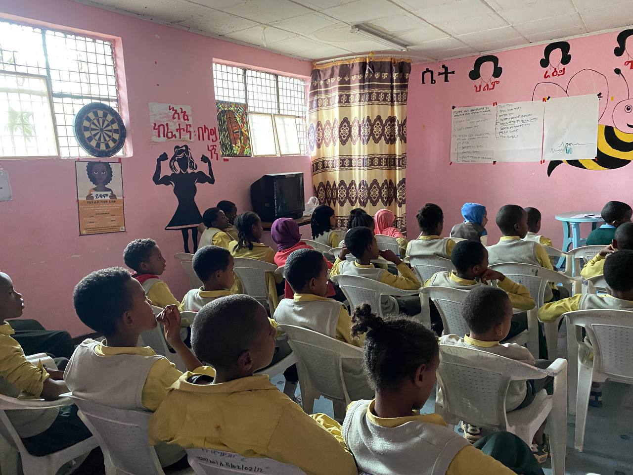 Vi ser en gjeng med elever i gul og beige skoleuniform i et rosa klasseom med tegninger på veggene. Inkludert en illustrasjon på veggen i sort av en dame som viser muskler. Vi ser elevene bakfra og ser at de kikker opp mot noen ark med skrift som er klistret på veggen foran dem.