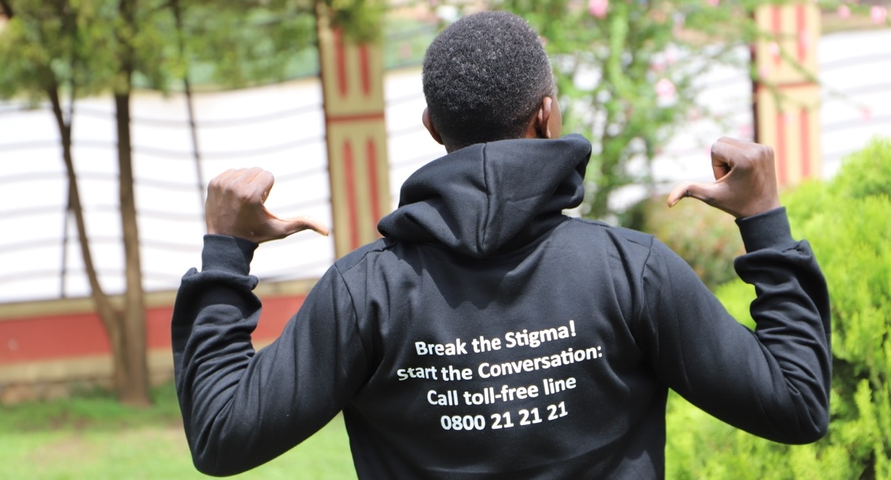 Mann med svart hettegenser står med ryggen mot kamera. På genseren står det i hvit skrift: "Break the stigma! Start the conversation: Call toll-free line" med et ugandisk telefonnummer under.