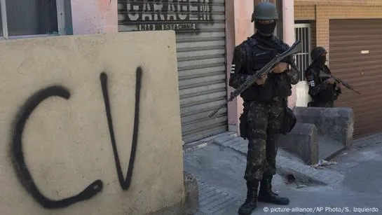 policial em frente a sigla do Comando Vermelho pixada em muro
