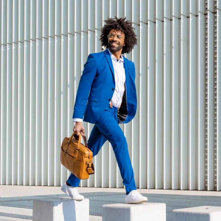 Ein Mann in einem auffälligen blauen Anzug schreitet dynamisch mit einer braunen Ledertasche vor einer modernen Fassade aus vertikalen Streben entlang.