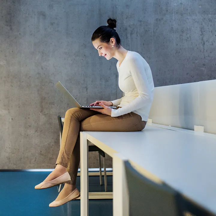 Eine Frau in heller Kleidung sitzt auf einem Tisch und arbeitet konzentriert an einem Laptop; der Hintergrund zeigt eine Betonwand.