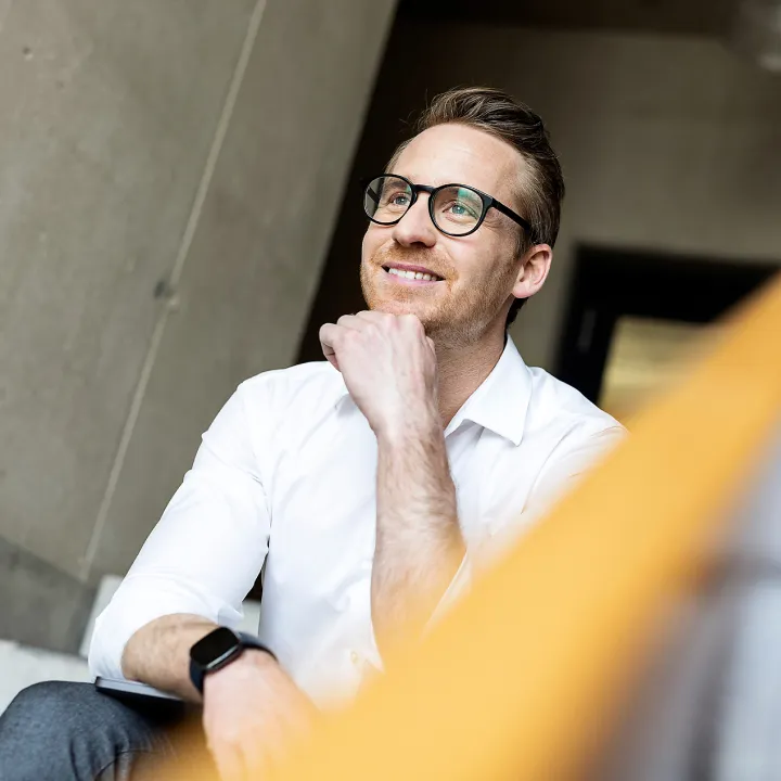 Ein Mann in einem weißen Hemd sitzt nachdenklich, stützt das Kinn auf die Hand und blickt zur Seite, im Hintergrund eine Betonwand.