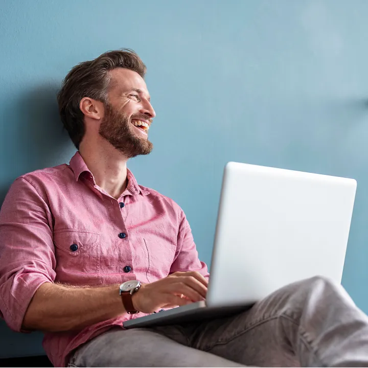 Ein Mann in einem rosa Hemd sitzt entspannt an einer blauen Wand und arbeitet mit einem Laptop auf dem Schoß.